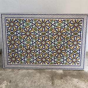 May include: A rectangular mosaic tabletop featuring an intricate geometric pattern of interlocking stars and lines in shades of deep blue, mustard yellow, and teal green, framed by a white and blue braided border on a white background.