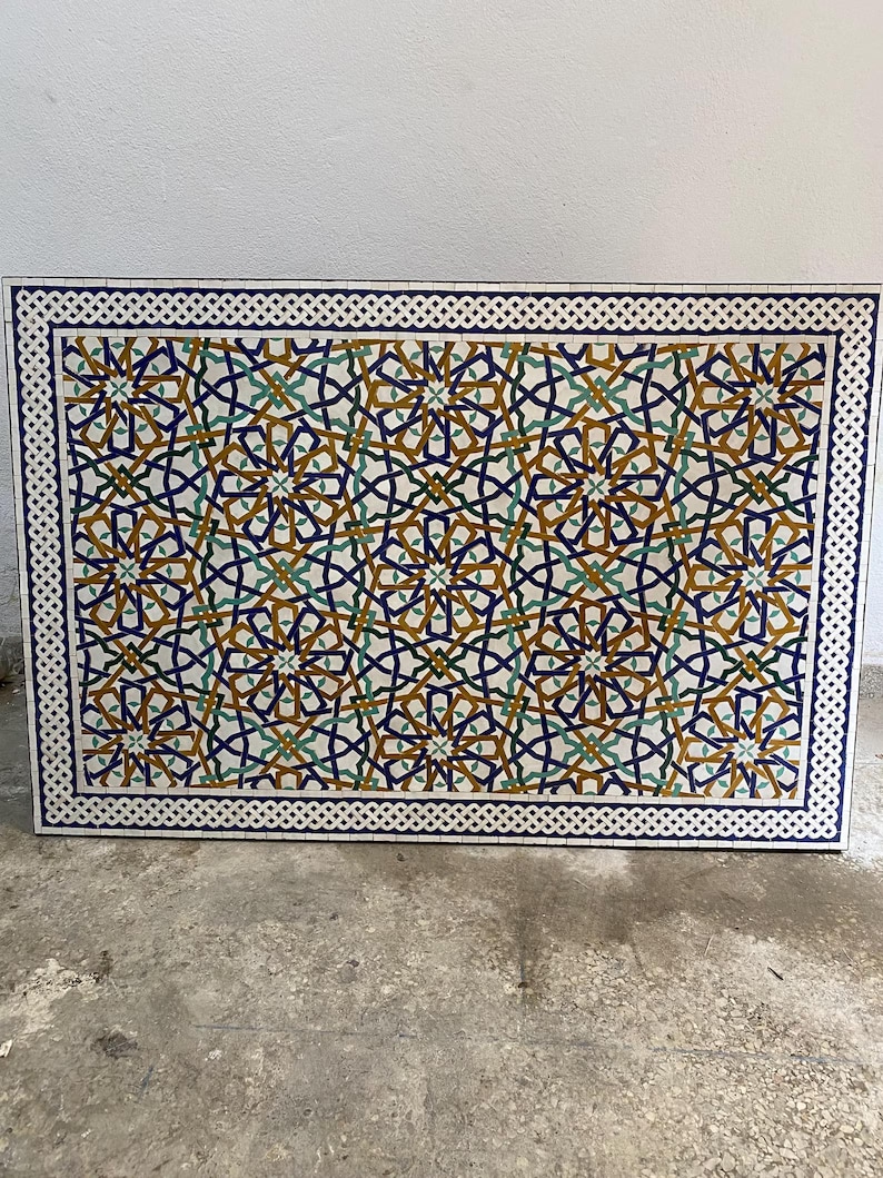 May include: A rectangular mosaic tabletop featuring an intricate geometric pattern of interlocking stars and lines in shades of deep blue, mustard yellow, and teal green, framed by a white and blue braided border on a white background.