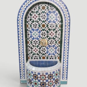 Può includere: Una fontana a muro ad arco caratterizzata da intricati motivi geometrici a mosaico nei toni del blu, verde, bianco e marrone. La struttura include un rubinetto centrale in ottone e una vasca semicircolare alla base, decorata con piastrelle.