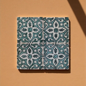 Op de afbeelding: Vier vierkante keramische tegels met een turquoise en wit bloemenpatroon. De tegels zijn samen gerangschikt en vormen een grotere vierkant. De achtergrond is effen perzik van kleur, met een schaduw van het zonlicht.