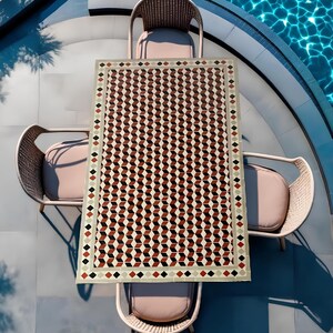May include: A square outdoor dining table with a geometric pattern of red, black, and white tiles. The table is surrounded by four wicker chairs with beige cushions. The table is set on a patio with a swimming pool in the background.