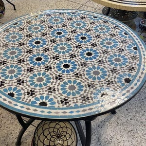 Peut inclure: Une table ronde avec un plateau en mosaïque présentant un motif géométrique répétitif dans des tons de bleu, blanc, gris et noir. La table possède un cadre en métal noir à quatre pieds et une étagère inférieure contenant un bol à motif linéaire.