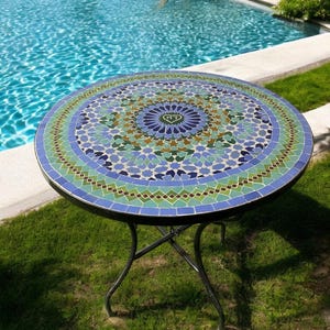 Könnte beinhalten: Ein runder Mosaiktisch mit einem geometrischen Muster in Blau, Grün und Braun. Der Tisch hat ein schwarzes Metallgestell und steht auf einem grünen Rasen.