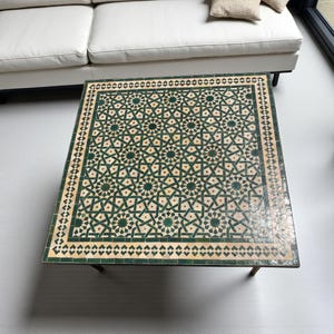 May include: Square coffee table with a geometric tile top in shades of green and beige. The intricate pattern features star and diamond shapes. The table has a dark metal frame and is positioned in front of a white sofa.