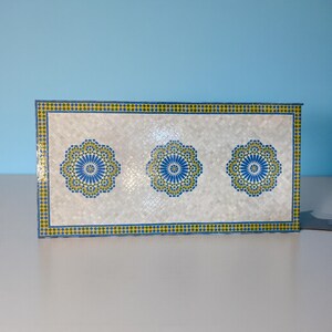 May include: A rectangular decorative box featuring three blue and yellow floral designs on a white background. The box is bordered with a blue and yellow diamond pattern. The background has a textured, woven appearance.