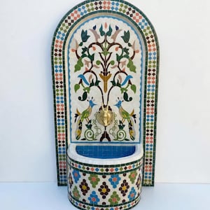 Puede incluir: Fuente de pared de mosaico con un diseño central de árbol y dos pavos reales. La fuente tiene una pila azul y un grifo dorado. El mosaico presenta una variedad de colores, incluyendo azul, verde y marrón.