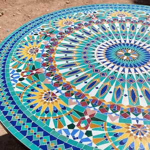 May include: A round mosaic tile table with a detailed geometric pattern in shades of blue, teal, yellow, and white. The table has a black metal frame and legs. The mosaic design features intricate shapes and patterns.