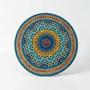 Op de afbeelding: Een rond tafelblad met een complex geometrisch mozaïekpatroon in tinten blauwgroen, mosterdgeel, donkerblauw en grijs. Het midden toont een klein bordeauxrood hartje met de letters MT, omringd door concentrische stervormige motieven.