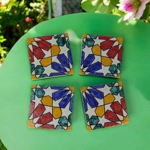 May include: Four square ceramic coasters with a geometric star pattern in red, blue, yellow, and green, outlined in black. The coasters are arranged on a green surface, with a blurred background of greenery and flowers.
