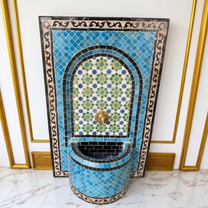 Könnte beinhalten: Dekorativer Wandbrunnen mit Mosaikfliesen-Design. Der Brunnen hat einen hellblauen Fliesen-Hintergrund mit einem zentralen bogenförmigen Design und einem Messing-Löwenkopf-Ausguss. Das Becken hat eine gebogene Form, ebenfalls blau gefliest.