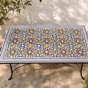 May include: A rectangular table featuring a mosaic tile top with a repeating geometric star pattern in shades of blue, yellow, green, and white. The table has a black metal frame with curved legs and a white border with a braided knot design.