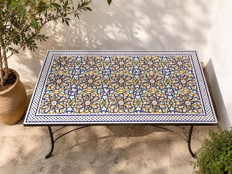 May include: A rectangular table featuring a mosaic tile top with a repeating geometric star pattern in shades of blue, yellow, green, and white. The table has a black metal frame with curved legs and a white border with a braided knot design.