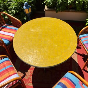 Pode incluir: Uma mesa redonda ao ar livre com um tampo de mosaico amarelo brilhante. A mesa é rodeada por quatro cadeiras de vime com almofadas listradas coloridas. A cena é montada em um tapete vermelho, com vegetação ao fundo.