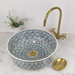 Peut inclure: Un lavabo en céramique décoratif avec un motif de vagues bleu et blanc. Un robinet doré et un porte-savon sont également visibles. Le lavabo est rond et possède un drain doré. Le robinet a des poignées en croix.