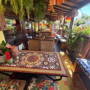 May include: A rectangular outdoor dining table with a colorful geometric mosaic tile top featuring red, green, and yellow patterns. The table is surrounded by wicker chairs with cushions, hanging pendant lights, and lush green hanging ferns.