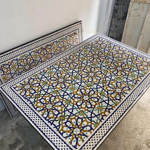 May include: A rectangular table with a mosaic tile top featuring an intricate geometric pattern in shades of blue, yellow, and green. The border consists of a white and black interlocking knot design, framing the complex star-shaped motifs.