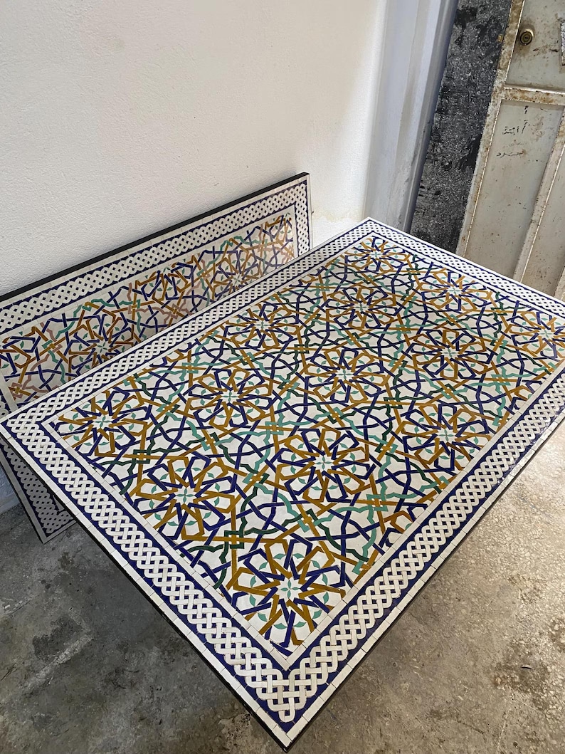 May include: A rectangular table with a mosaic tile top featuring an intricate geometric pattern in shades of blue, yellow, and green. The border consists of a white and black interlocking knot design, framing the complex star-shaped motifs.