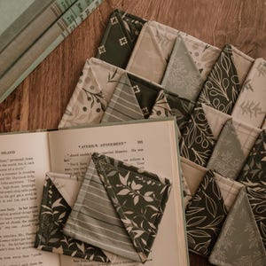 May include: A collection of square fabric book corners featuring various patterns in shades of sage green, olive, cream, and white. The designs include botanical leaf prints, subtle stripes, and geometric motifs, all stitched with visible quilted seams.