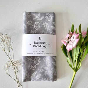 May include: A gray beeswax bread bag with a white floral pattern. The bag is labeled "Beeswax Bread Bag" and "handmade with love". It is reusable, washable, and sustainable.