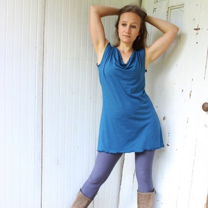 May include: A teal-colored sleeveless dress with a cowl neck and a scalloped hemline. The dress is paired with lavender leggings and worn with tan-colored boots. The model is leaning against a white wooden door.