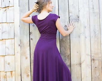 May include: A woman wearing a long, purple maxi dress with short sleeves. The dress has a fitted bodice and a flowing skirt. She is standing in front of a wooden wall.