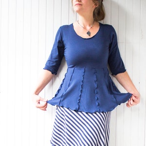 Op de afbeelding: Een vrouw draagt een blauwe top met een gerimpelde zoom en een wit-blauwe gestreepte rok. Ze draagt ​​bruine laarzen en een zilveren ketting met een hanger.