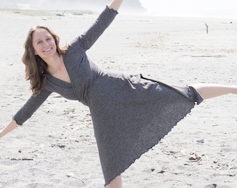 May include: A woman in a gray dress is standing on a sandy beach with her leg extended to the side. She is smiling and looking at the camera.