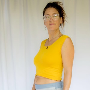May include: A yellow crop top and a light blue and white striped skirt. The top is sleeveless and has a scoop neck. The skirt has a wrap design. The person is wearing glasses and a brown leather bracelet.