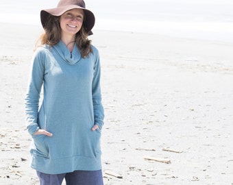 May include: A woman wearing a light blue tunic top with a brown wide-brimmed hat, standing on a sandy beach.