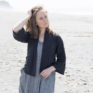 May include: A woman wearing a dark blue jacket over a grey dress, standing on a sandy beach.