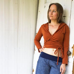 May include: A woman wearing a rust-colored wrap top with a hood and a navy blue waistband. The top is tied at the waist and the woman is standing in front of a white wooden door.