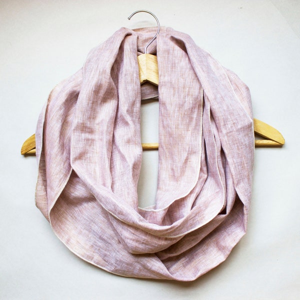 Peut inclure: Une écharpe infinie mauve clair avec une texture tissée visible, drapée en plusieurs boucles sur un cintre en bois. Le tissu présente un mélange subtil de fibres rose pâle et beige, créant un aspect sourd et naturel sur toute la pièce.