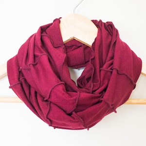 Puede incluir: Una bufanda infinita color burdeos con un borde rizado. La bufanda está colgada de una percha de madera.