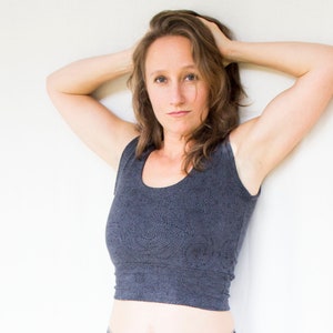 May include: A woman with long brown hair is wearing a dark gray sleeveless crop top with a subtle pattern. She is standing in front of a white wall with her arms raised above her head.