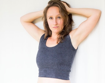 May include: A woman with long brown hair is wearing a dark gray sleeveless crop top with a subtle pattern. She is standing in front of a white wall with her arms raised above her head.