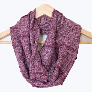 Puede incluir: Una bufanda infinita color burdeos con un patrón de punto texturizado, colgada de una percha de madera.
