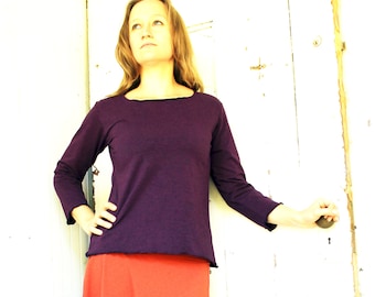 Puede incluir: Una mujer con una camisa de manga larga morada con cuello redondo y una falda naranja brillante. Está de pie frente a una puerta de madera blanca.