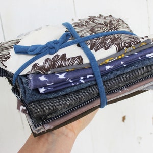 Puede incluir: Una pila de recortes de tela en varios tonos de azul, marr&oacute;n y blanco, atados con una banda el&aacute;stica azul. Los recortes de tela incluyen un estampado floral, un estampado de p&aacute;jaros y un estampado a rayas.