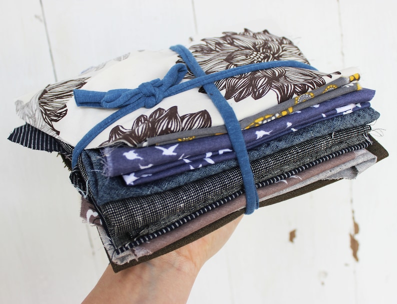 Puede incluir: Una pila de recortes de tela en varios tonos de azul, marr&oacute;n y blanco, atados con una banda el&aacute;stica azul. Los recortes de tela incluyen un estampado floral, un estampado de p&aacute;jaros y un estampado a rayas.