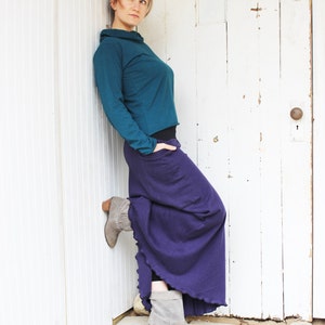 May include: A woman wearing a teal long-sleeved shirt and a long purple skirt. She is standing in front of a white door with a white trim. She is wearing grey ankle boots.