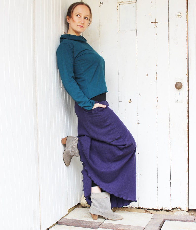May include: A woman wearing a teal long-sleeved shirt and a long purple skirt. She is standing in front of a white door with a white trim. She is wearing grey ankle boots.