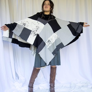 Könnte beinhalten: Ein Patchwork-Poncho in Schwarz-, Weiß- und Grautönen. Der Poncho hat einen Rollkragen und ein geschichtetes Design. Das Model trägt ein graues Kleid und braune Stiefel. Das Design des Ponchos besteht aus verschiedenen Stoffquadraten.