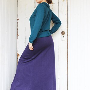 May include: A woman wearing a teal long-sleeved shirt and a long purple skirt. She is standing in front of a white door with a door knob.