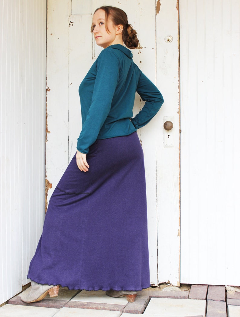 May include: A woman wearing a teal long-sleeved shirt and a long purple skirt. She is standing in front of a white door with a door knob.