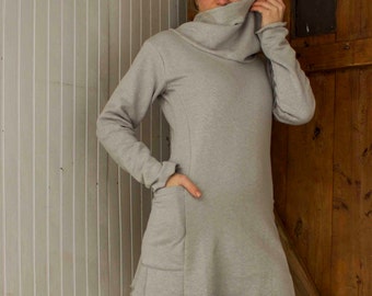 May include: A gray cowl neck sweater dress with long sleeves and a pocket on the front. The dress has a ruffled hem and is worn with black leggings.