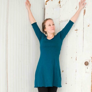 May include: A woman wearing a teal blue long-sleeved dress with a scoop neck and a slightly flared hem. She is standing with her arms raised above her head, looking up.