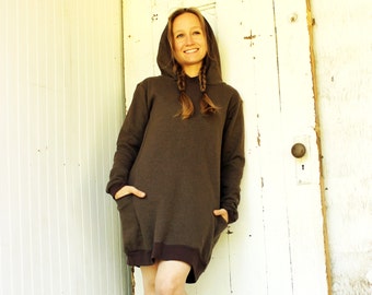 May include: A brown hooded sweatshirt dress with long sleeves and a relaxed fit. The model is wearing the dress with her hair in two braids.