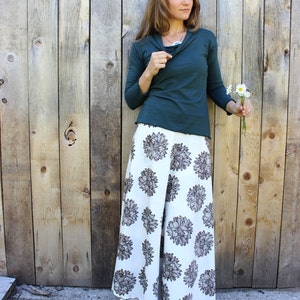 Puede incluir: Una mujer con una camisa de manga larga de color turquesa y una falda blanca con estampado floral marr&oacute;n. Est&aacute; de pie frente a una pared de madera y sostiene un ramo de flores blancas.