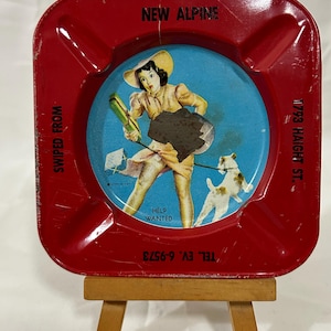 May include: Red metal ashtray with a blue and white image of a woman in a hat holding a dog leash and a dog. The text "NEW ALPINE" is at the top of the ashtray. The text "SWIPED FROM" is on the left side of the ashtray. The text "HELP WANTED" is in the center of the ashtray. The text "TEL. EV. 6-9573" is on the bottom left side of the ashtray. The text "1793 HAIGHT ST." is on the right side of the ashtray.