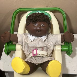 Könnte beinhalten: Eine Vintage-Puppe der Cabbage Patch Kids mit dunkler Haut und braunen Haaren, die in einem cremefarbenen Hochstuhl mit einem grünen Griff sitzt. Die Puppe trägt ein beiges Outfit mit weißem Kragen und gelben Schuhen. Ein weißer Gürtel mit einer rosa Schnalle sichert die Puppe.